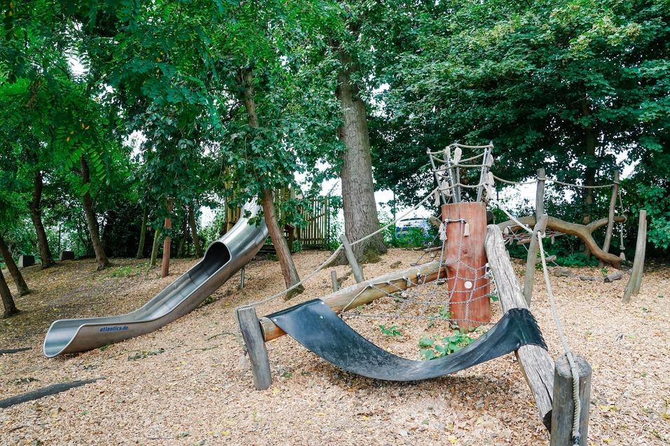 Spielplatz mit einer Metallrutsche, Klettergerüst aus Holz und weichem Bodenbelag aus Rindenmulch, umgeben von Bäumen.