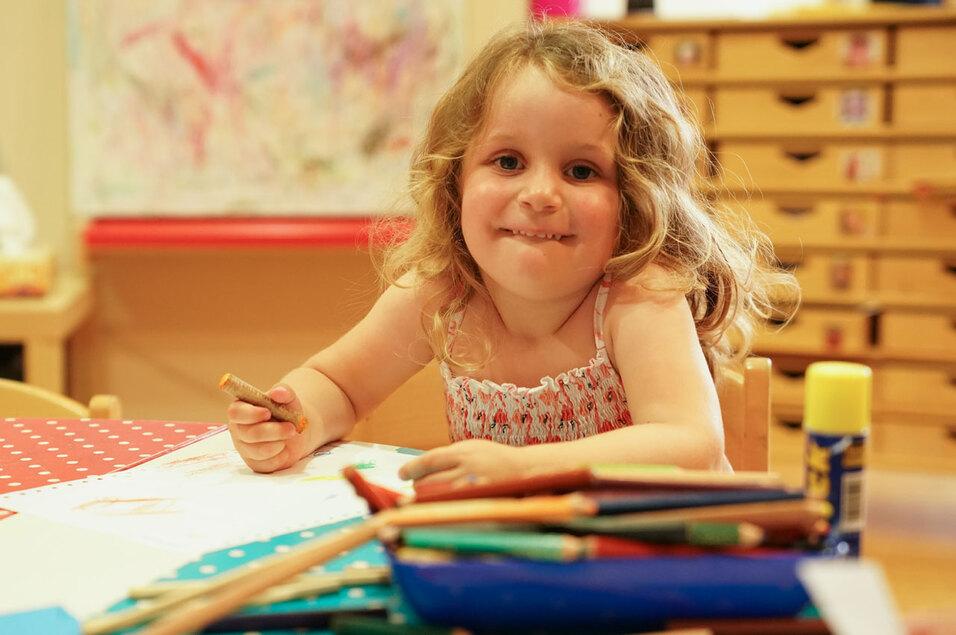 Ein kleines Mädchen mit lockigem Haar sitzt an einem Tisch und zeichnet mit Buntstiften auf Papier.