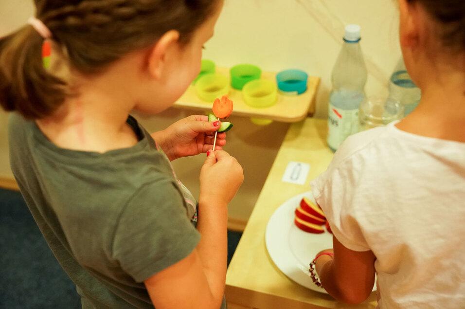 Kinder gestalten Obstspieße mit einer Karottenblüte und Gurkenscheibe in einer kreativen Spielumgebung.