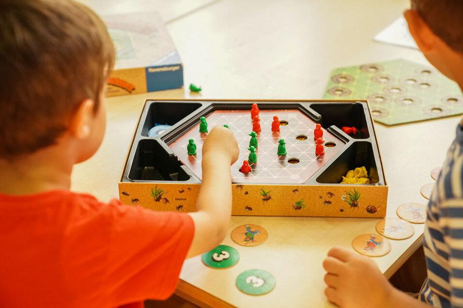 Zwei Kinder spielen ein Brettspiel mit bunten Figuren auf einem hexagonalen Spielfeld. Spielmarken liegen daneben.