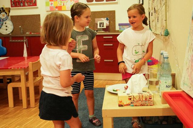 Drei Kinder spielen in einem bunten Raum, während sie Snacks auf einem Tisch teilen und miteinander lachen.