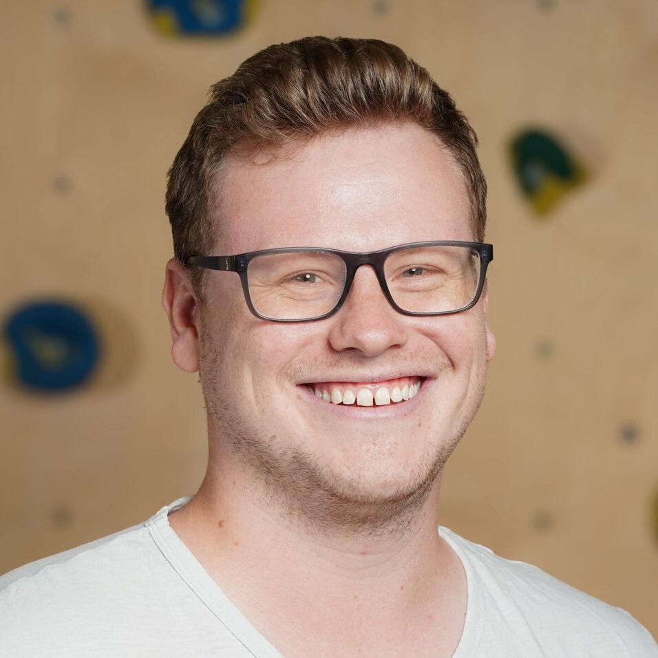 Lächelnder Mann mit Brille vor einer Kletterwand aus Holz mit bunten Griffen.