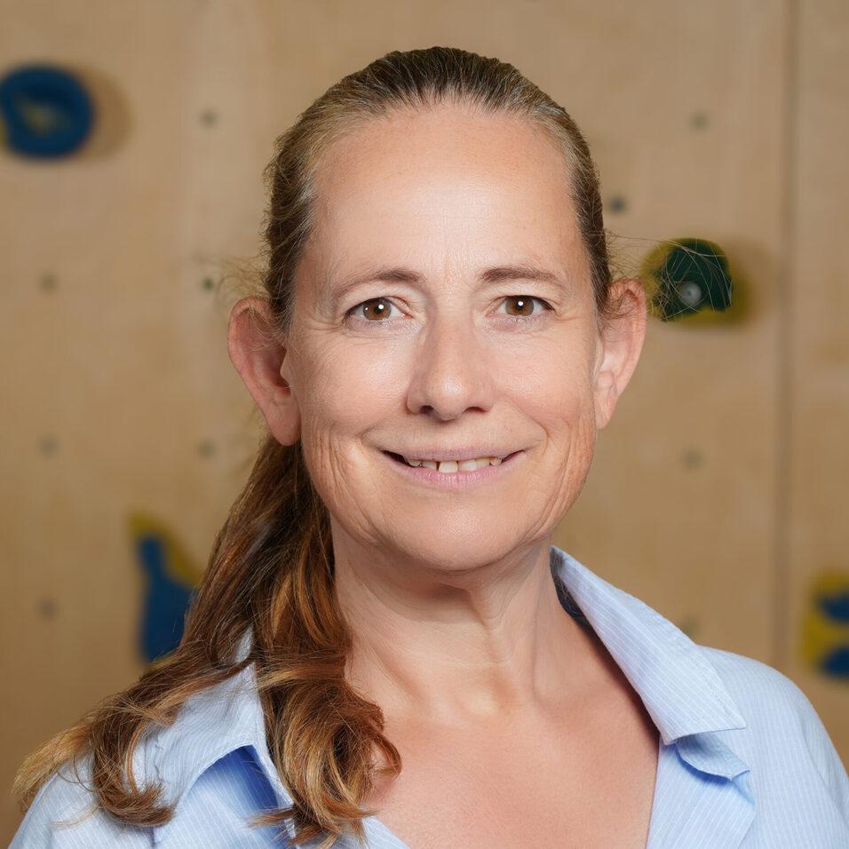 Portrait einer lächelnden Frau mit langen, braunen Haaren, getragen in einem blauen Hemd, vor einer Kletterwand.