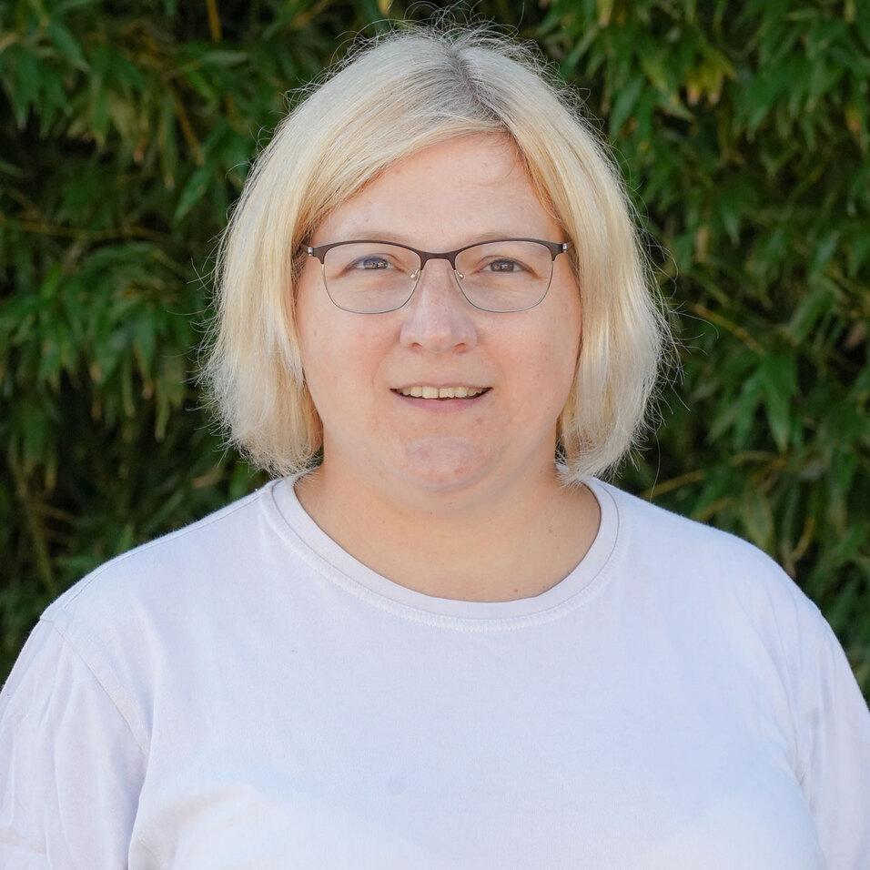 Porträt einer blonden Frau mit Brille, die vor einer grünen Pflanzenwand steht und lächelt.