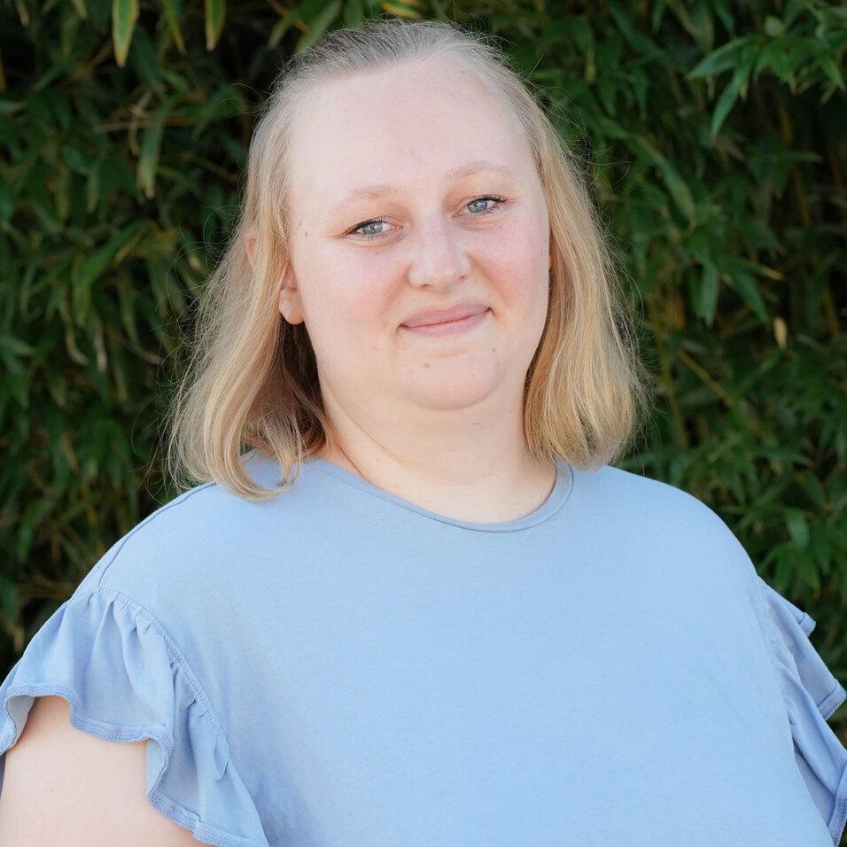 Porträt einer lächelnden Frau mit blonden, schulterlangen Haaren, getragen in einem hellblauen Oberteil vor grünem Hintergrund.